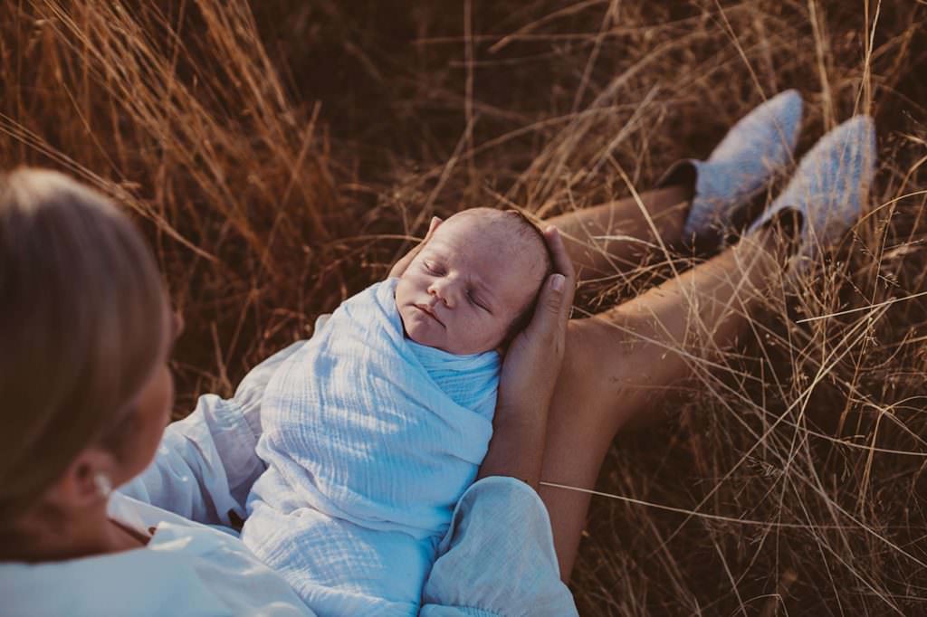 newborn baby that is wrapped in white cradled by his mother whilst sitting in golden grass