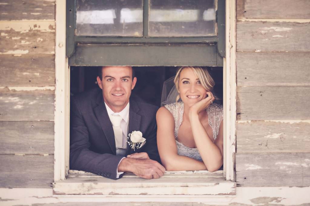 a bride and groom hangout the window of an old vintage house