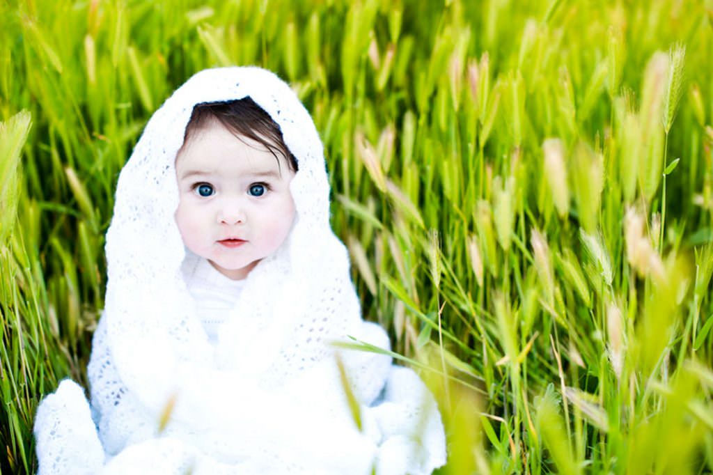 a todler with dark eyes ina vibrant green grass she is wrapped in a white baby blanket