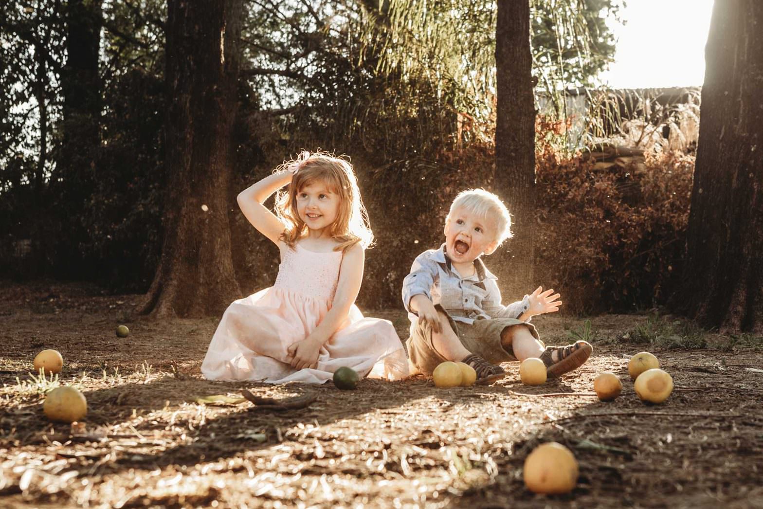 two kids sitting on a dirt road at The Pavillion. The sun is setting in the background. The kids are laughing & throwing fruit