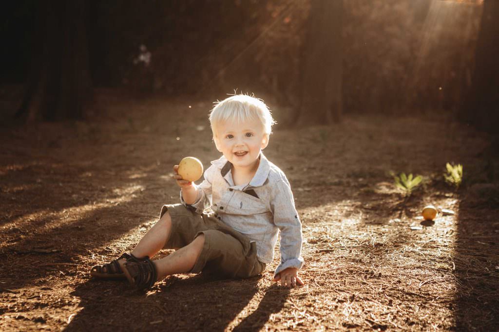 a boy holds a yellow peice of fruit with the suns rays streaming in the background. He has blonde hair & smiles at his Tamworth family photographer Karissa Tuckwell