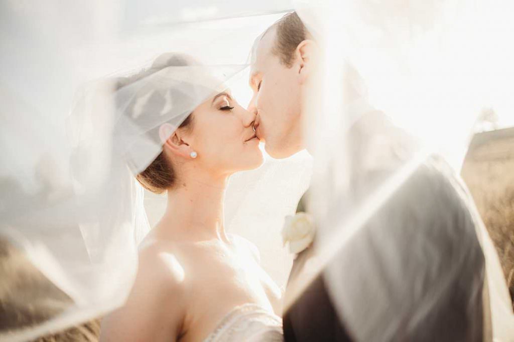 bride and groom kiss under the veil as they sunsets behind her veil