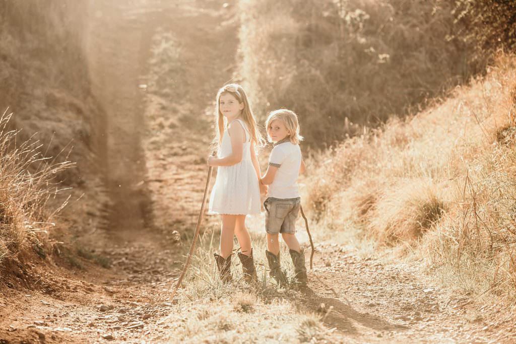 two blonde headed siblings with sticks in their hands wearing cowboy boots look around at their Tamworth family portrait photographer as the golden sun sets in the background