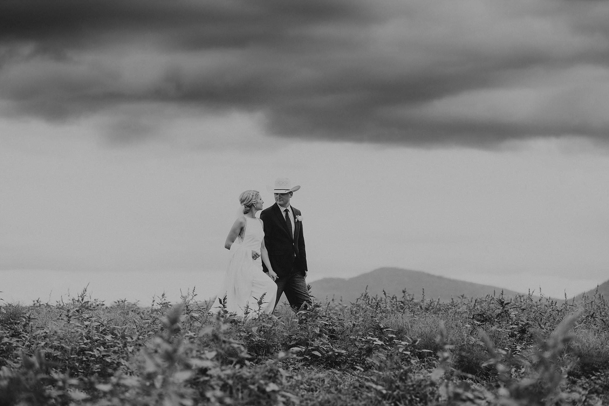 black and white photo the tamwoprth wedding photographer has captured the bride and groom with a cowboy hat walking with his bride in long grass.The bride is wearing a simple straight white wedding dress as she holds onto her husbands hand and her train. there is dark storm clouds