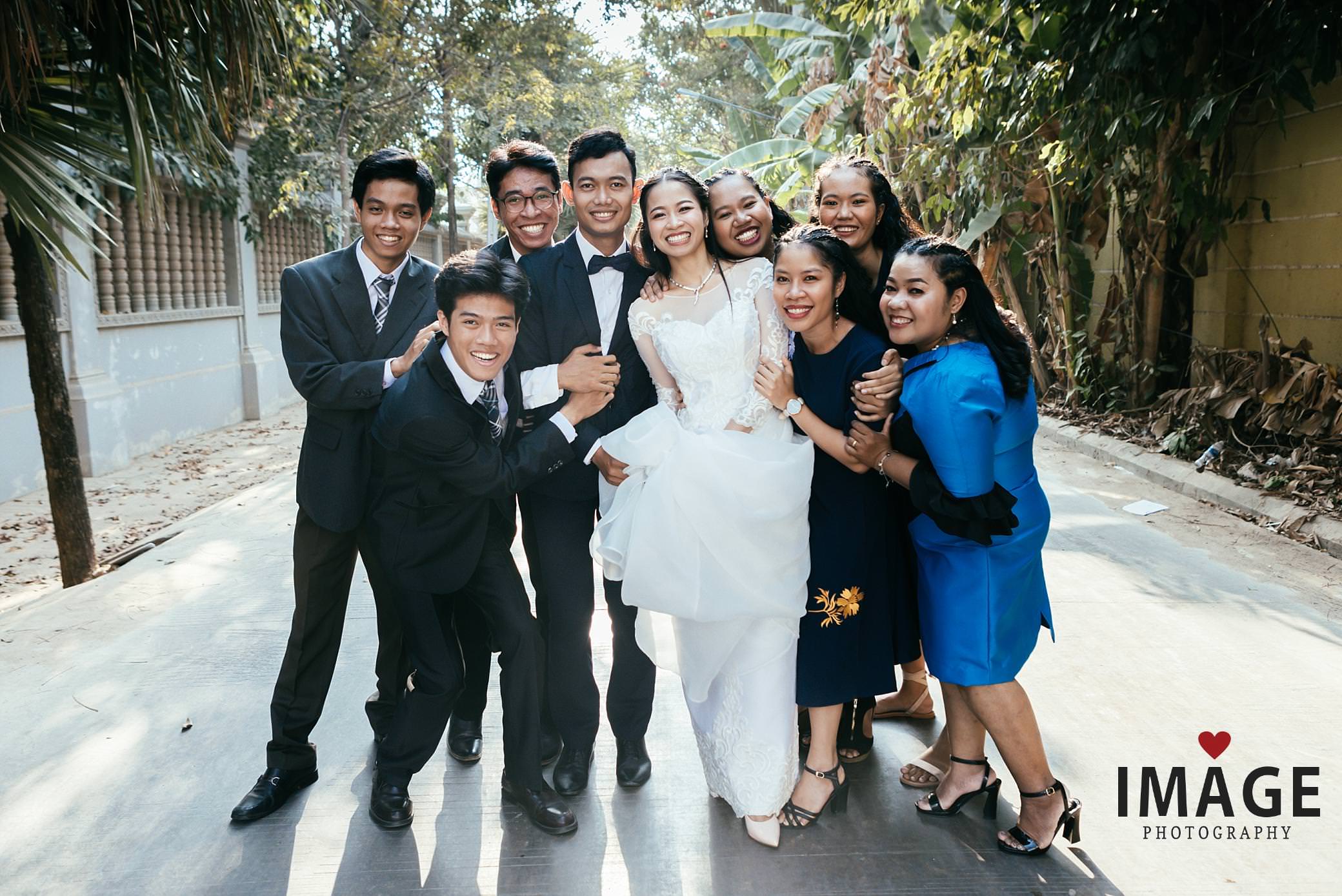 a bridal party hug the cambodian bride and groom in a tree lined road way