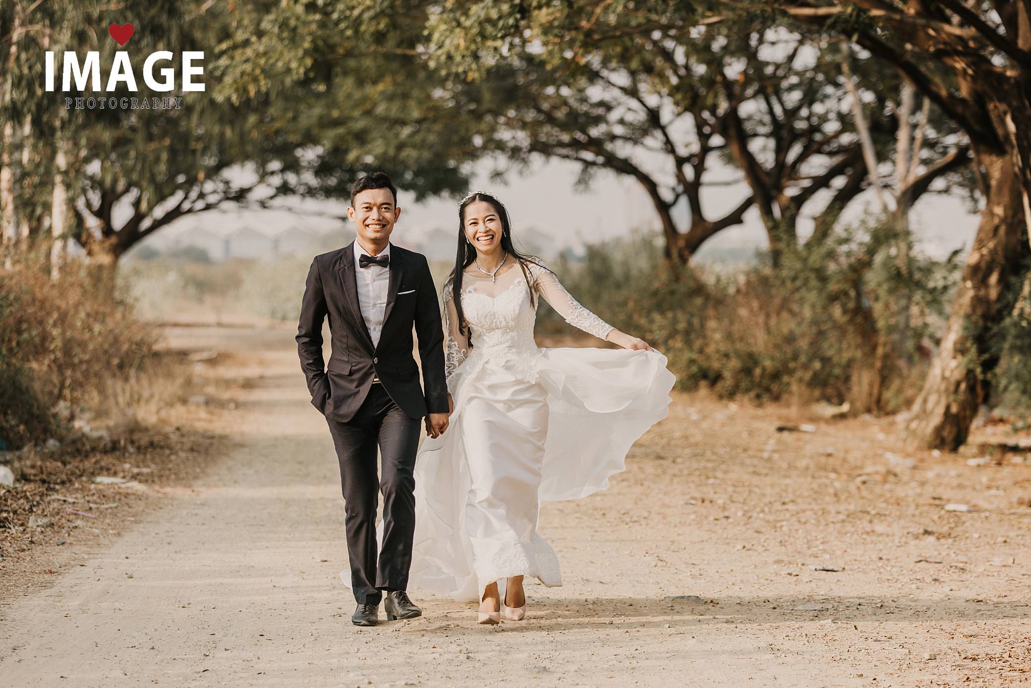 a tree lined road way with a bride and groom walk holding hands as she hold her white lace dress they are laughing on a road in cambodia
