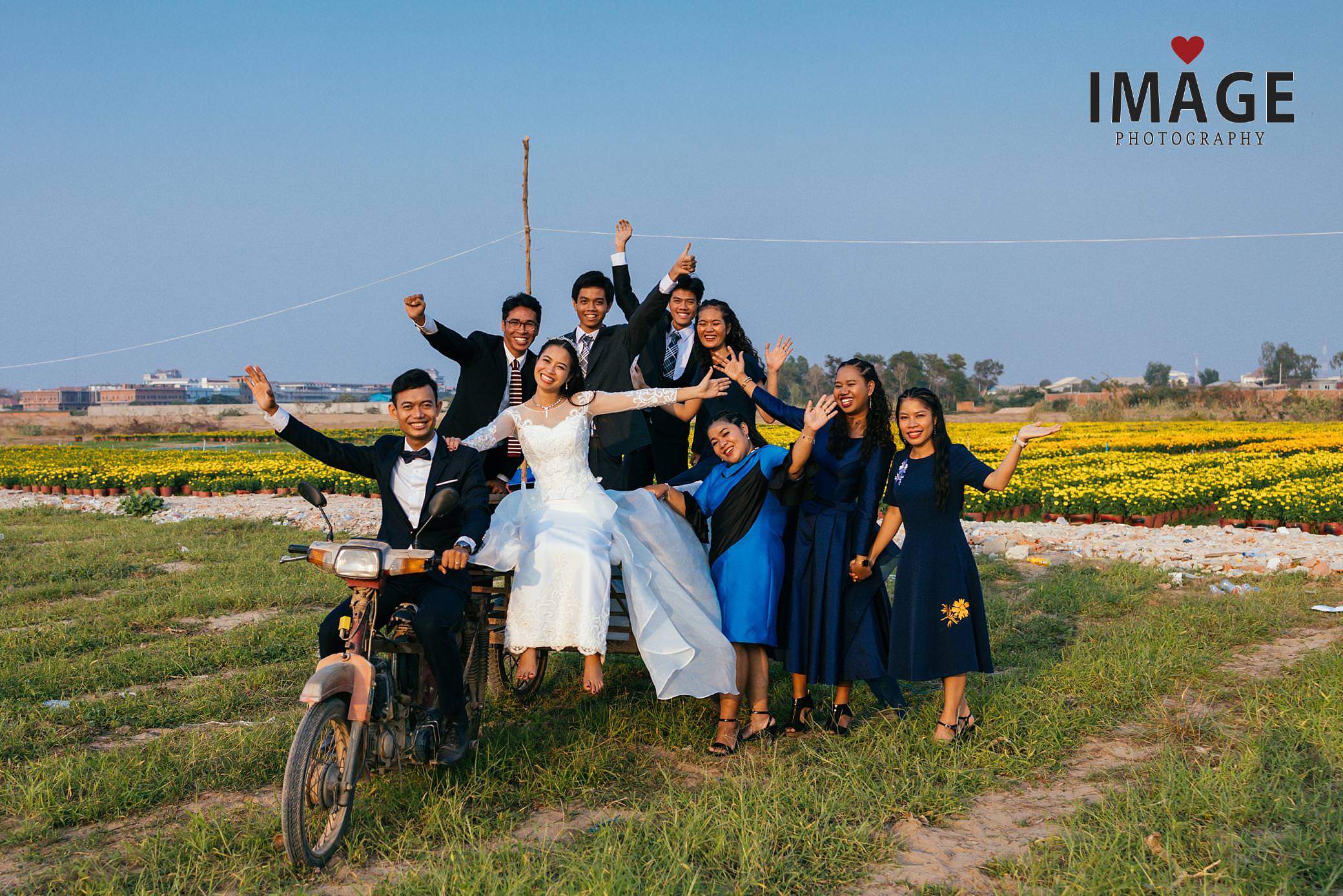 a cambodian bridal party poses with their friends ona cambodian motobike with yellow flowers in the background