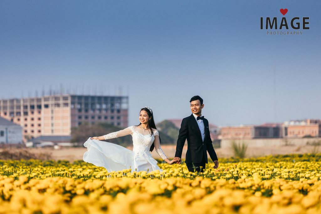 a bride holds her dress as she is led by her groom in a field of yellow flowers on the outskirts of phnom penh