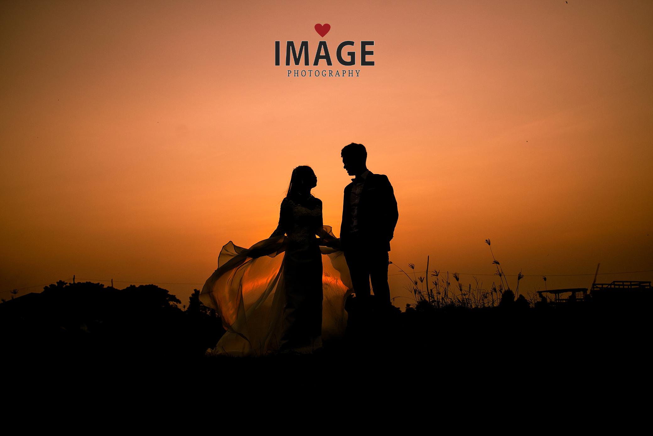 a phnom penh sunset is golden red with a groom & bride as a background with her dress blowing in the wind
