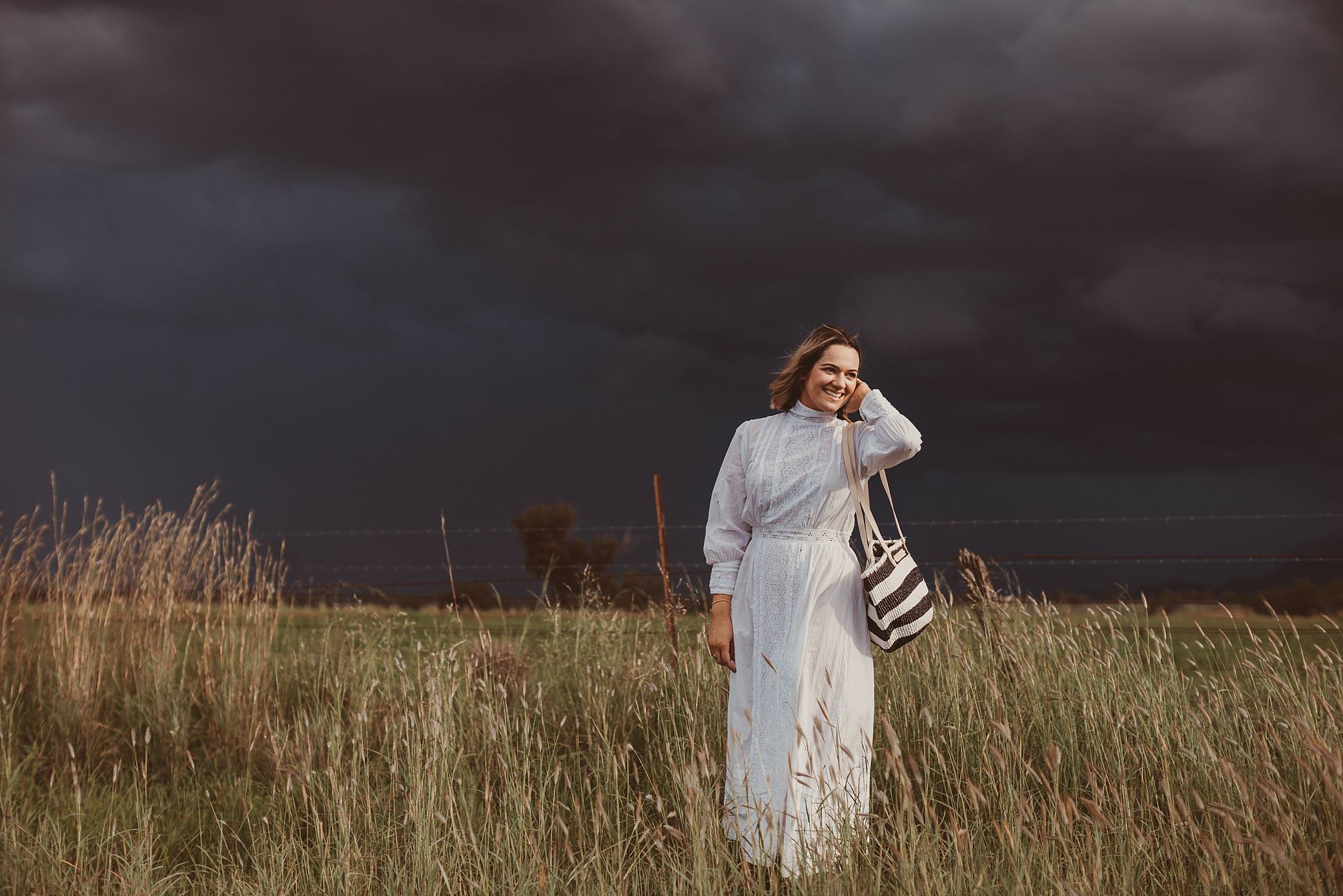 woman in a white vintage dress has her wrattan bag over her shoulder in country Australia as storm clouds roll in. she is tucking her hair behind her head