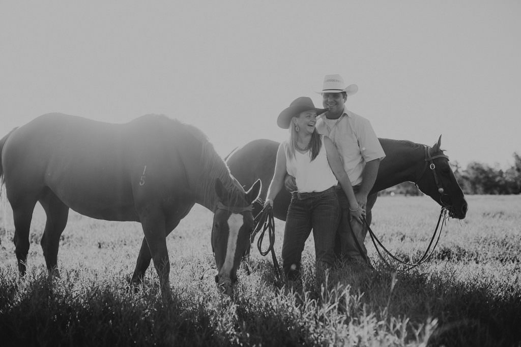black and white photo of a newly engaged couple hold onto each other. as the horses eat grass in the paddock