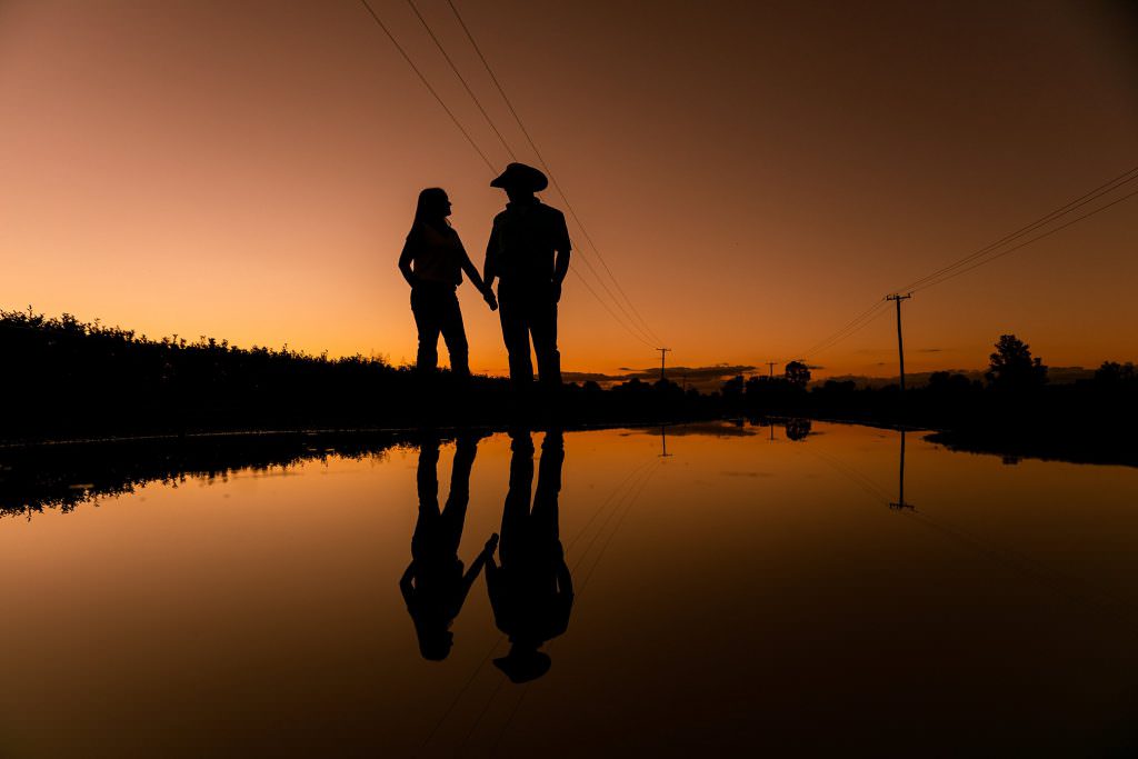 silhouette of a tamworth engaged couple holding hands as the sun goes down they are standing near a puddle with reflections and power poles the sky is orange