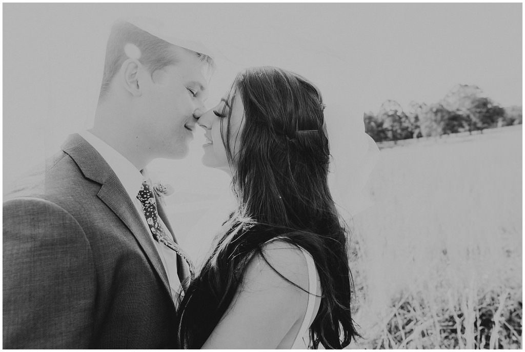 black and white photograph as a couple nearly kiss with smiles on the faces as the sun shines through her veil