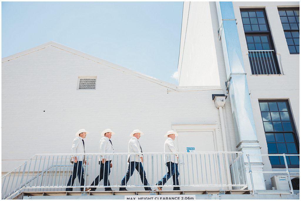 walking against a steel staircase with blue sky groomsmen in jackets and hats