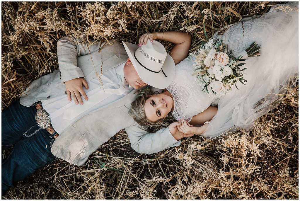 amongst a wheat crop a bride and groom lay down as the groom with a hat on looks at his bride & the bride wears a white lace dress