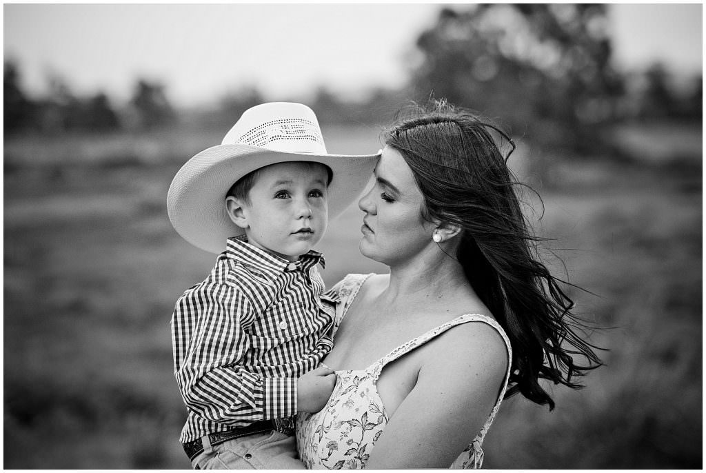 pure black and white photo of an innocent mum & son holding. Her hair is blown by the wind. He is looking to the Tamworth sky in his white cowboy hat