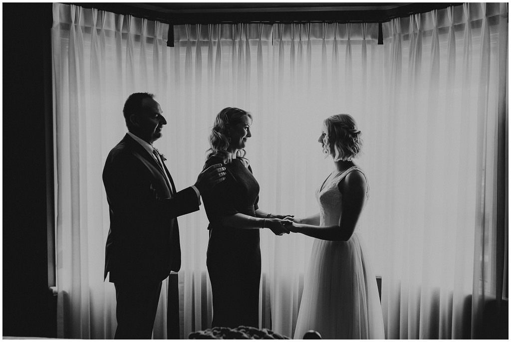 black and white image of a mum & dad holding thier daughters hand who is a bride as she gets ready in front of lace curtains