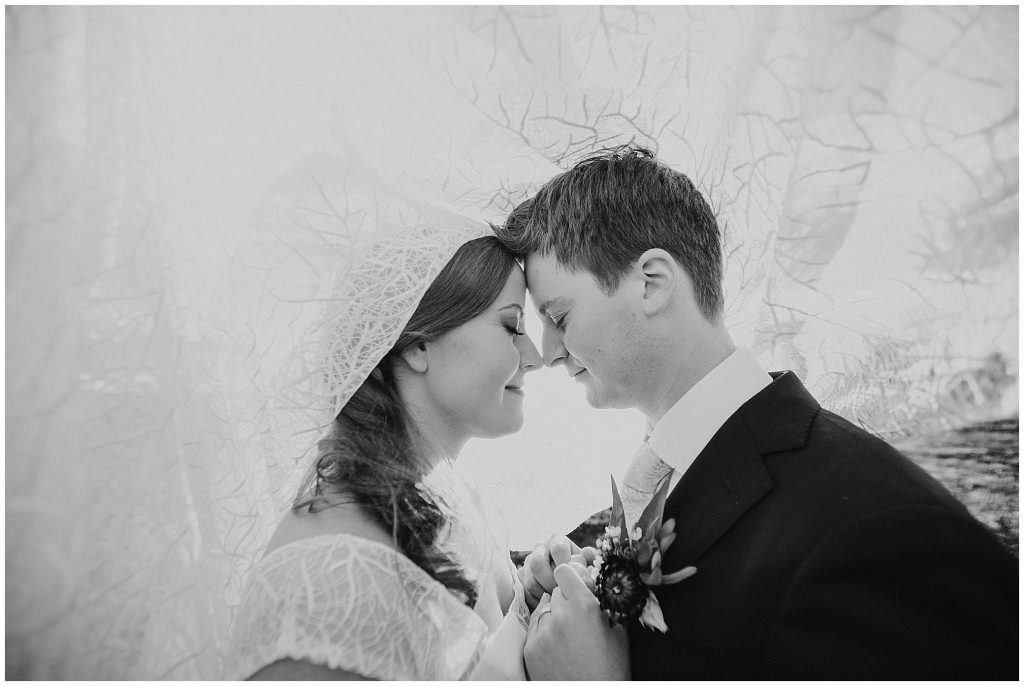 under a brides veil leaves and lace a bride and groom hug in a black and white photograph