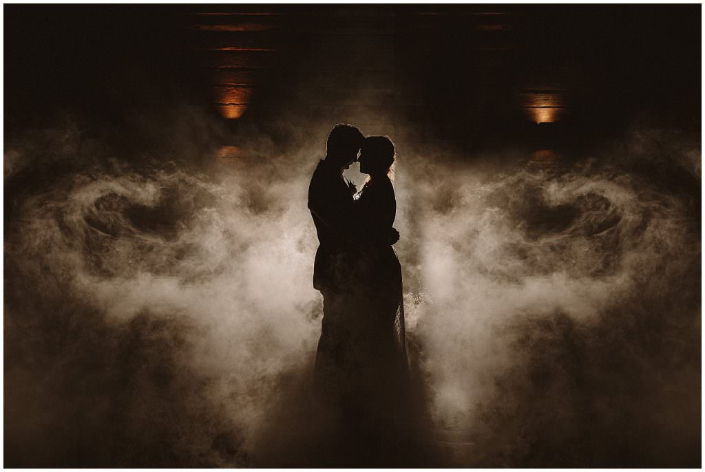 a bride and groom hold each other close at the entrance of Goonoo Goonoo Station, Tamworth NSW in the dark as fog swirls around them they are lit by lights