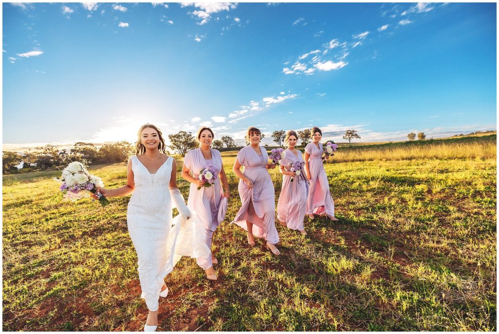 bride tribe walks holding flowers in purple bridesmaids dresses as the sun sets behind them