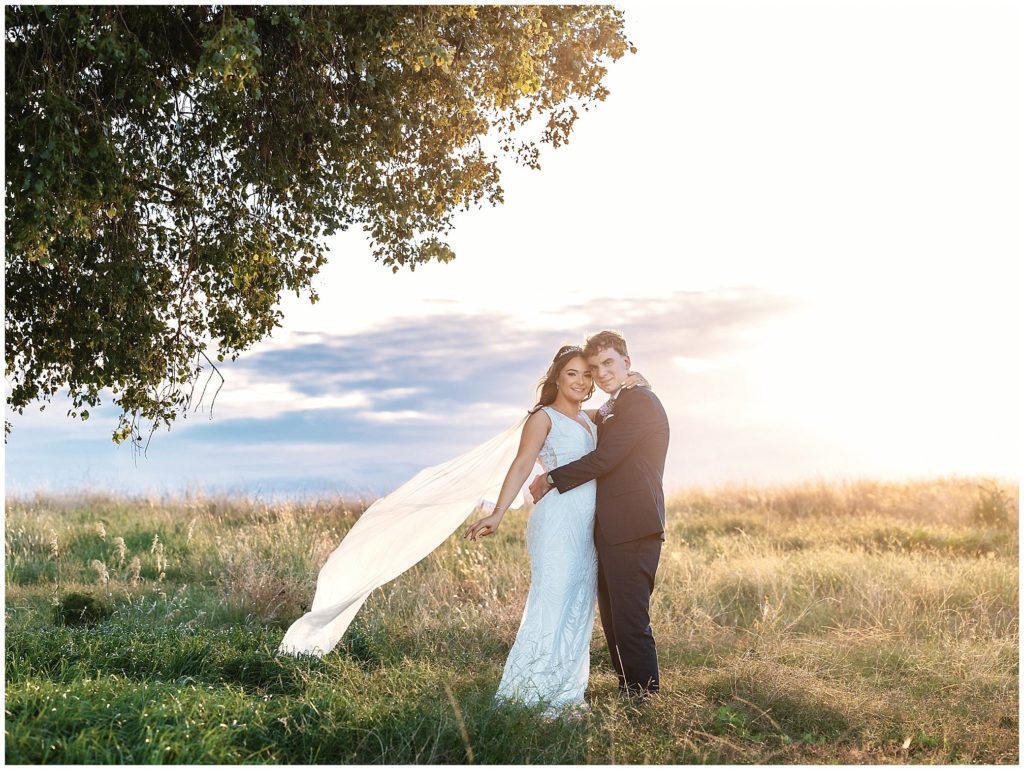 as the brides veil blows in the wind a couple hold onto each other as the sunsets. their is a moore creek tree next to them