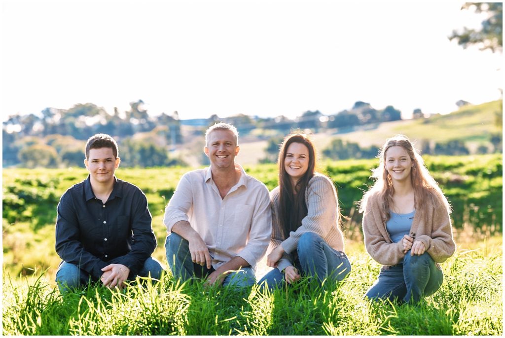 sit in the green grass a family of 4 smile as the sun sets