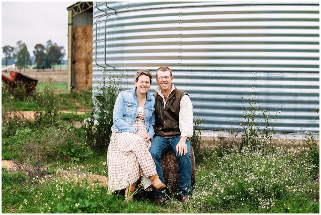 husband and wife sit amongst wild flowers with a rain water tank as a background