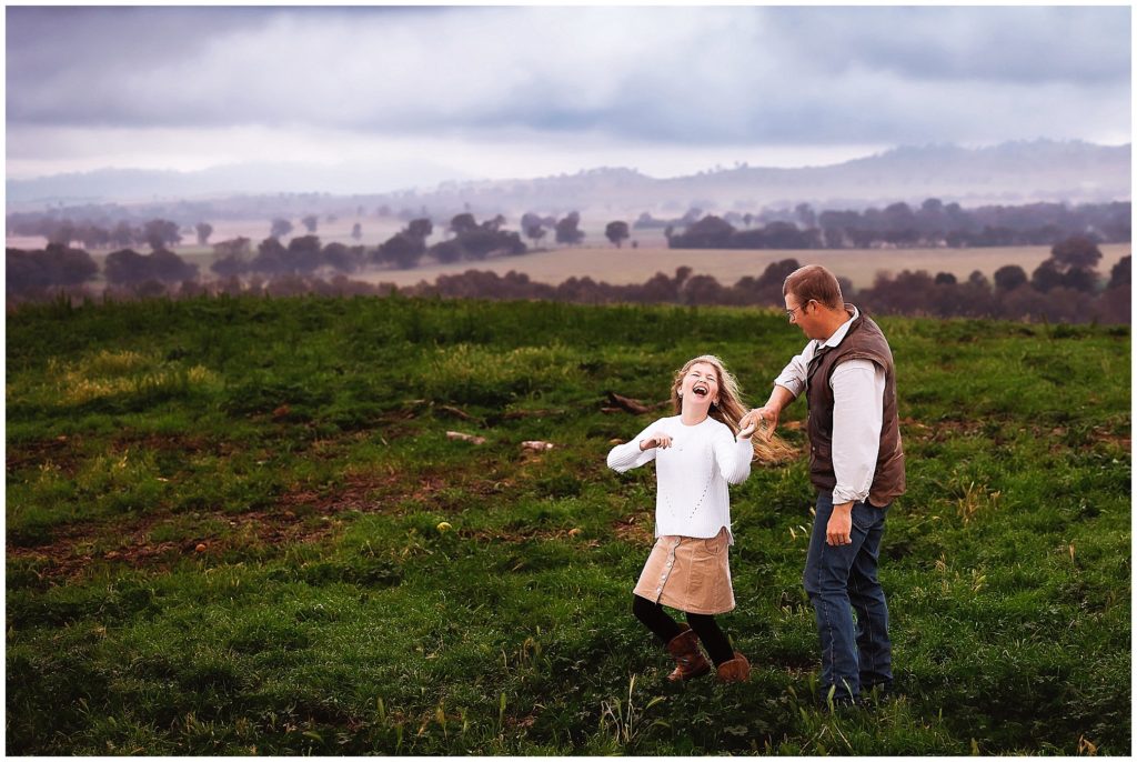 a dad spins his daughter who is in a brown skirt white shirt on top of a hill as the morning fog clears she laughs naturally at her dad in Cowra NSw