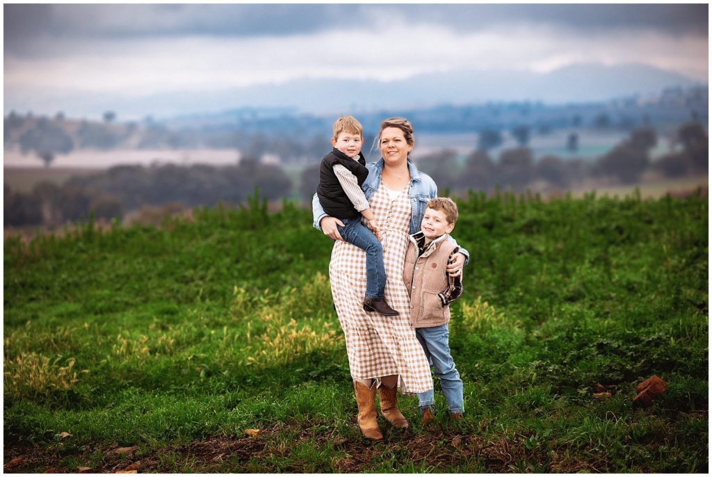 a mum and her two boys stand on a hills with storm clouds gree grass she hold one boy on her hip