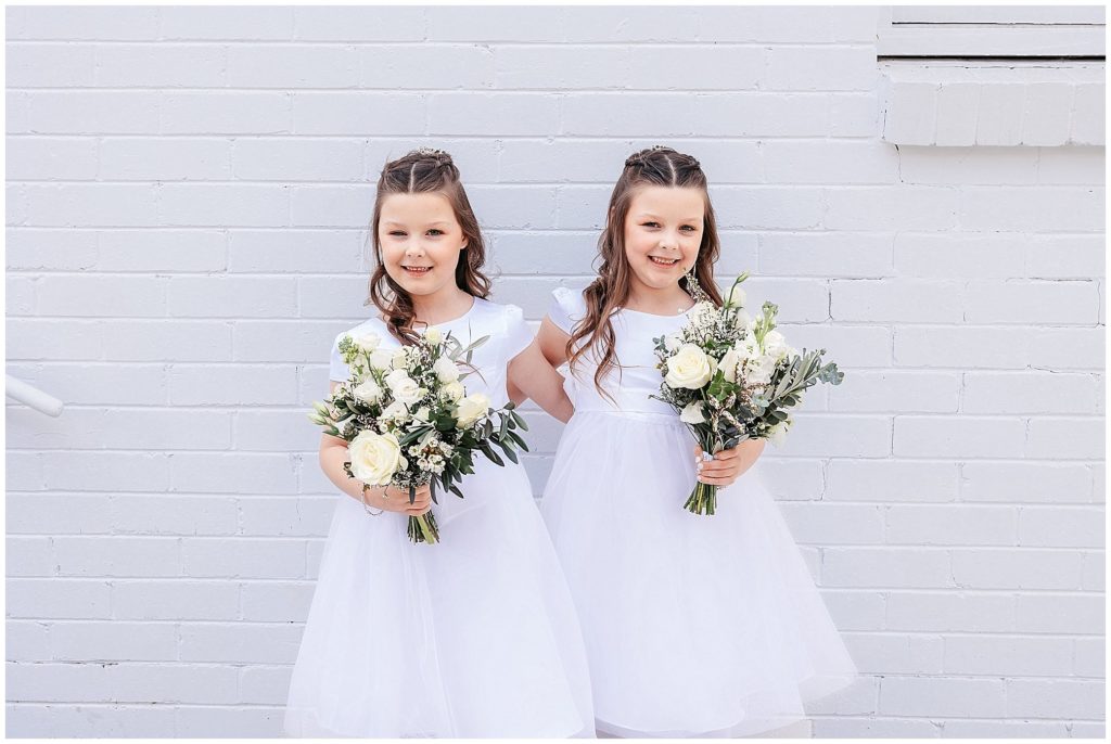 twin girls who wear white and white flowers against a white wall for a wedding