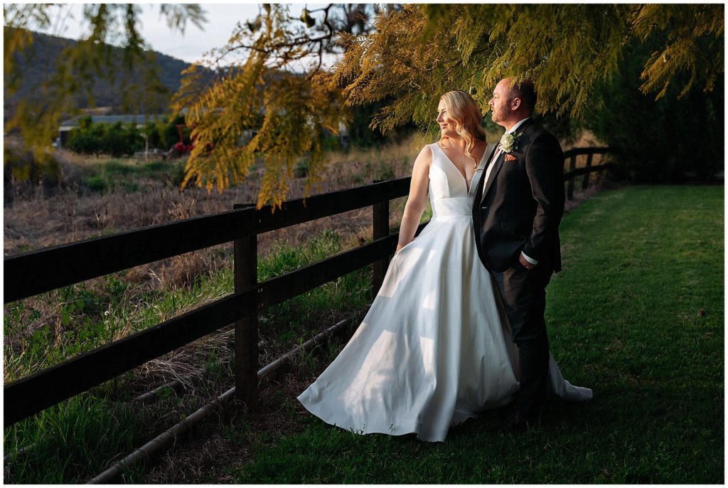 a bride with pockets in her white dress is held by her husband under a tree as they look at a Tamworth sunset near the Pavillion