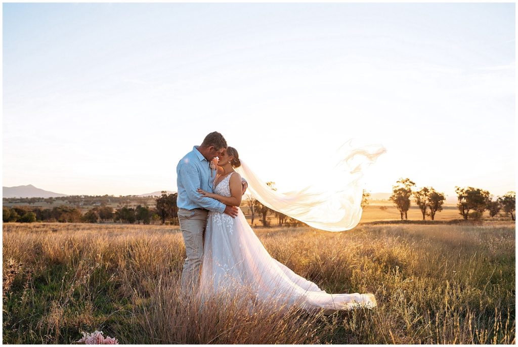 in the long grass on top of the hill at Dathem park wedding reception venue as a couple hug and almost kiss as the veil blows in the wind get photos as the sun sets