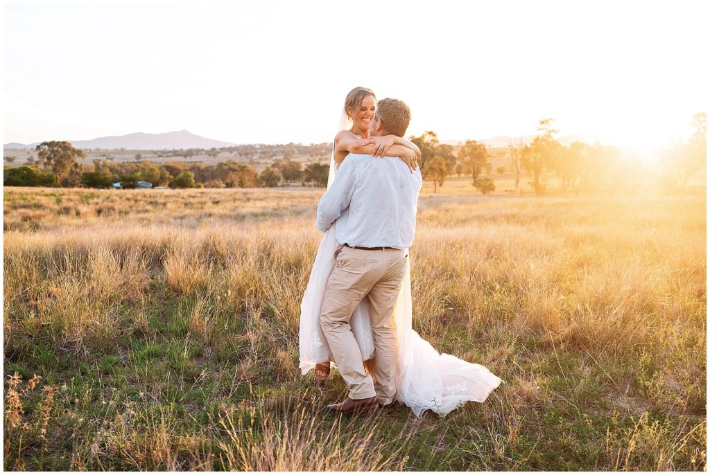 in the long grass on top of the hill at Dathem park wedding reception venue as a couple hug and almost kiss as the a bride and groom spin as they laugh as the sun sets over the Tamworth hills