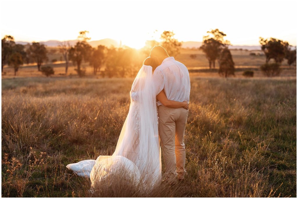 in the long grass on top of the hill at Dathem park wedding reception venue as a couple hug and almost kiss as the a bride and groom hug as the sun sets over the Tamworth hills