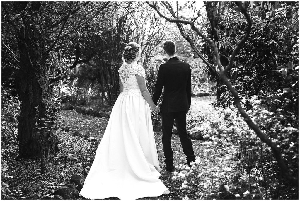 black and white of a bride and groom she has a lace back as they hold hands at Rock Abbey Gardens near Armidale nsw