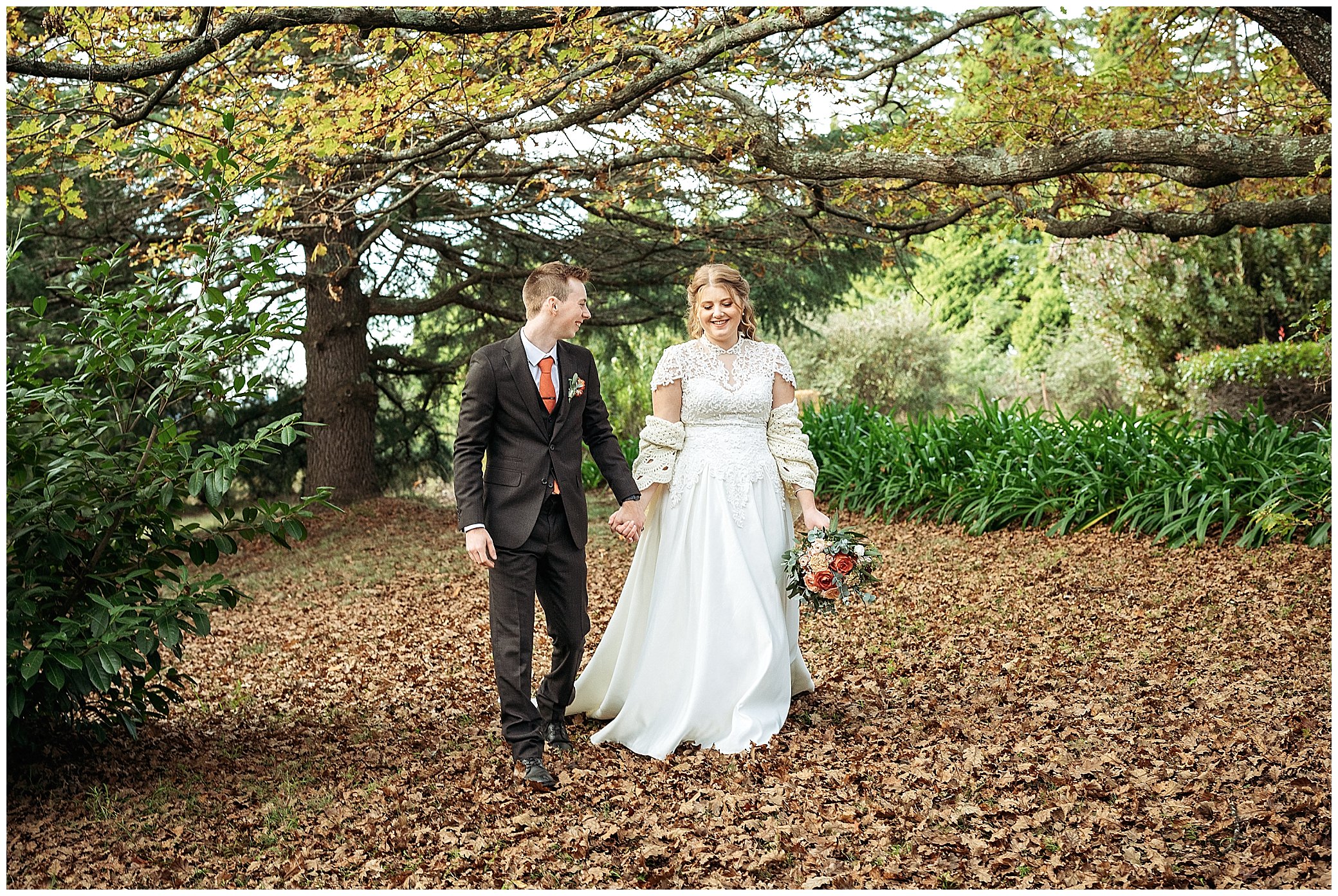 a bride wears a shawl knitted by her mother as she walks hand in hand with her husband under a big tree as she hold a bouquet made by Rosemerin florists