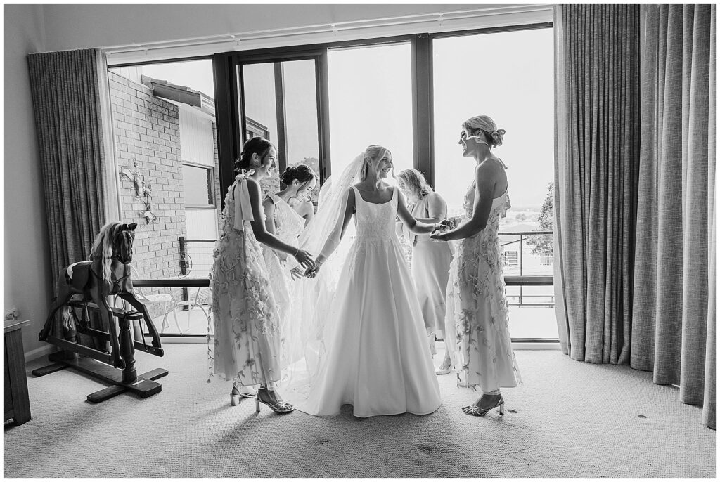black and white photo of a bride and her bridesmaid as they help get her ready she is holding her bridesmaids hands