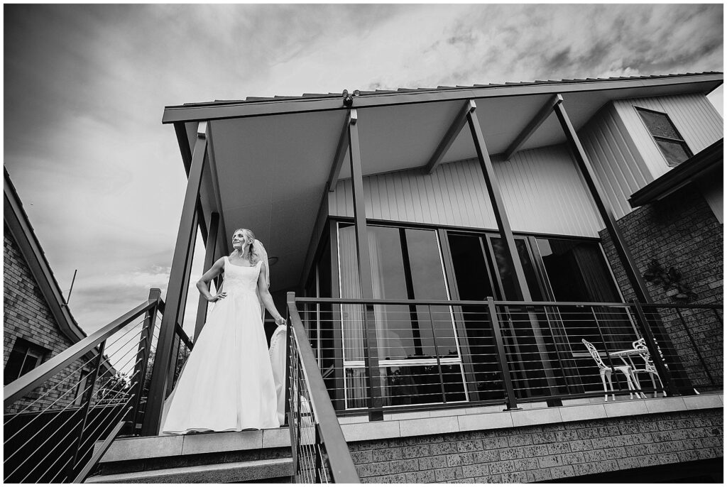 Black and white photo of a bride out the front of her childhood In Tamworth East NSW holds her dress and home smiles