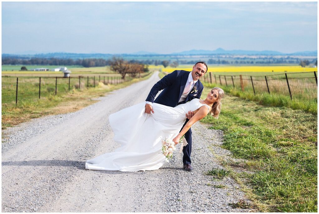 walking on a country road near Attunga nsw a couple dip each other as the groom laughs huge their is a canola crop in the background
