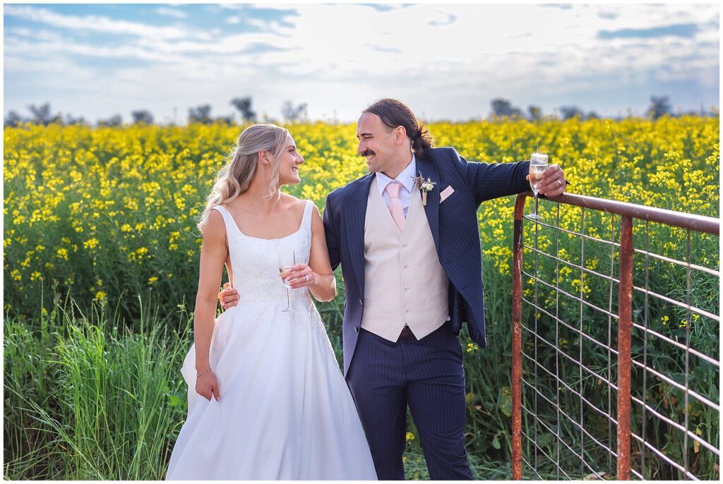 a bride and groom lean on a country fense as they share a drink together amongst a yellow canola crop Near Tamworth