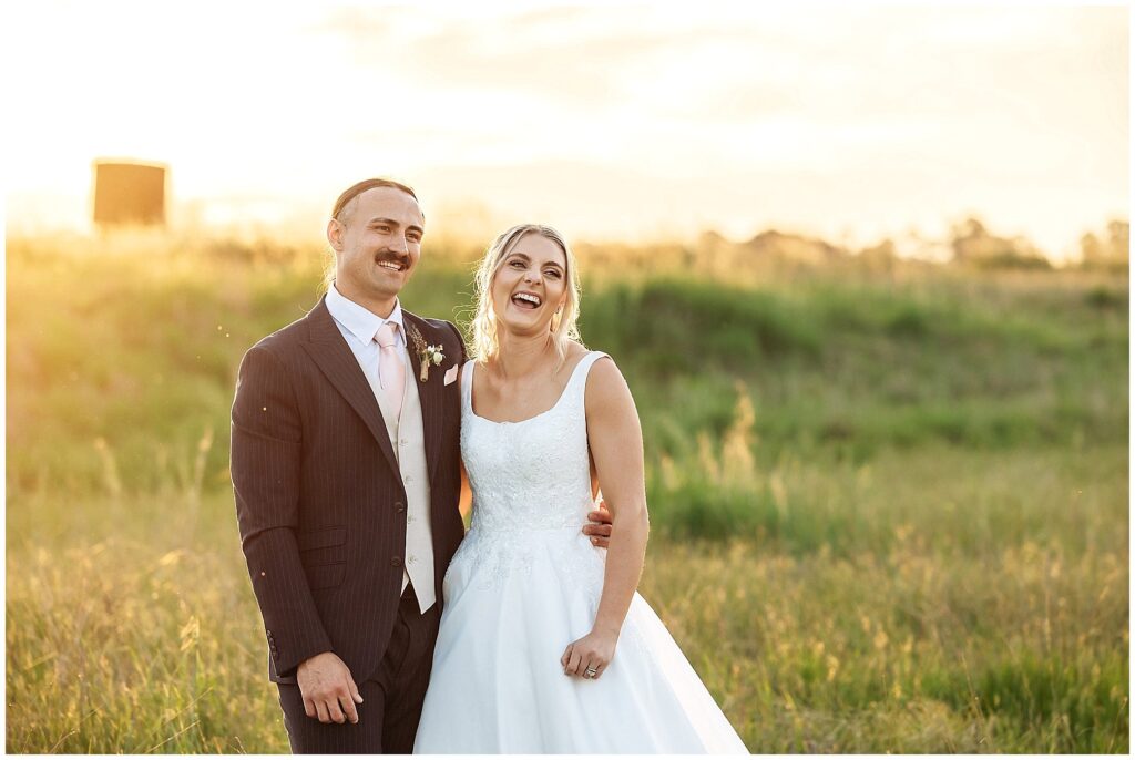 belly laughs as a newly married couple hold onto each other as the sun sets in the background