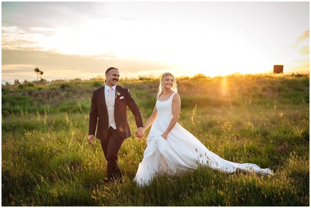 walking in the long golden grass for a bride and grooms holding hands as her dress drags as the sunsets over Tangarratta